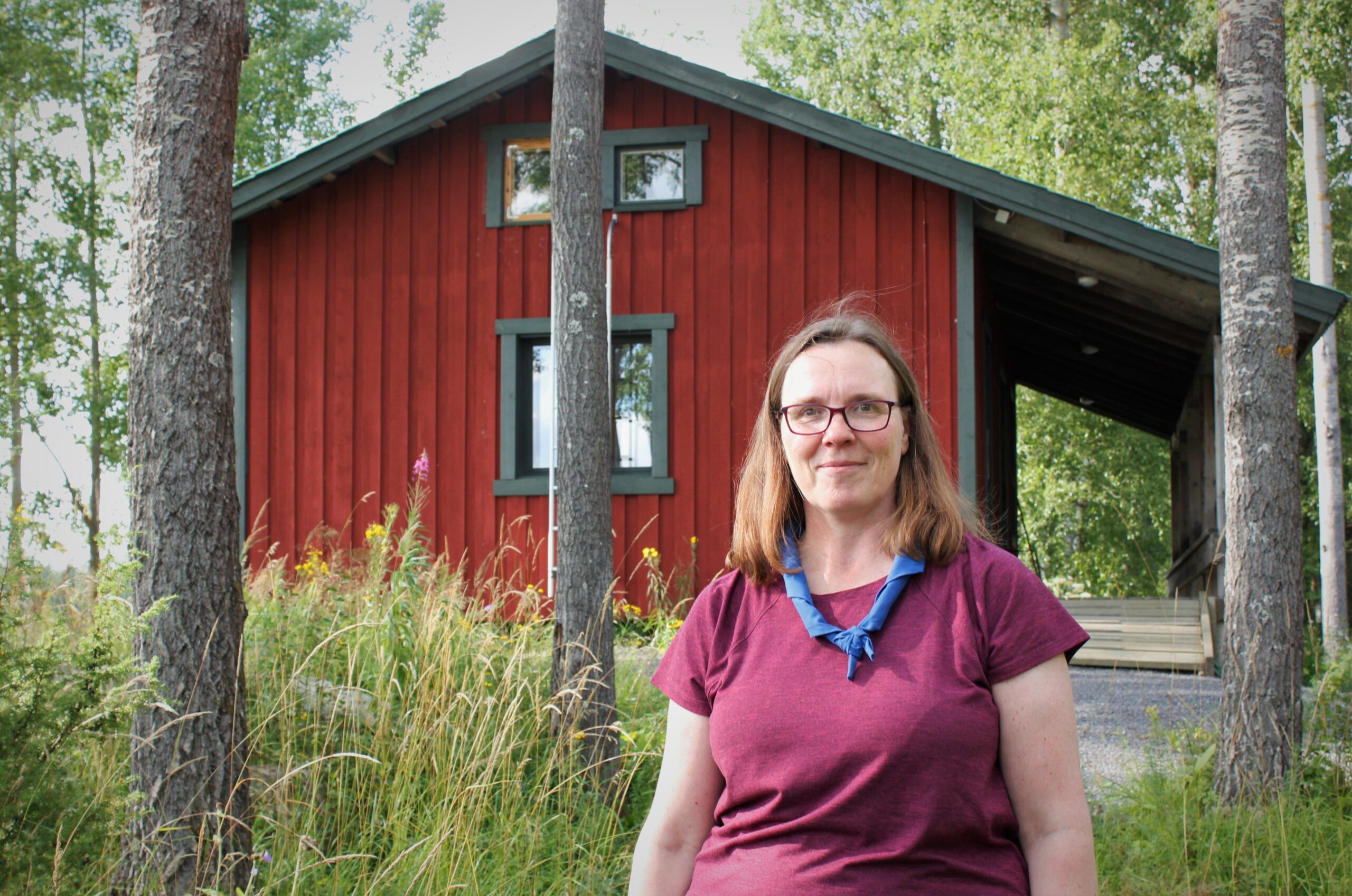 henkilö partiohuivissa punaisen kämpän edessä, puita